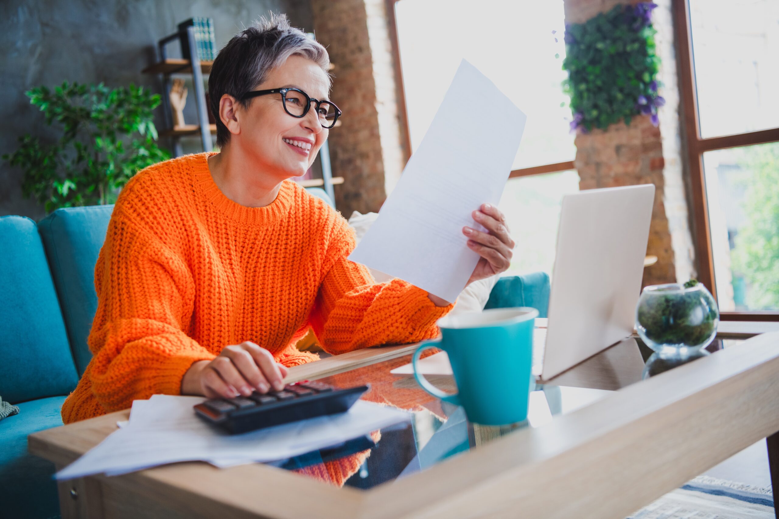 A person with short gray hair and glasses sits on a couch, smiling while looking at a document in one hand and using a calculator with the other. A laptop, papers, and a turquoise mug are on the table. The room is well-lit with large windows.