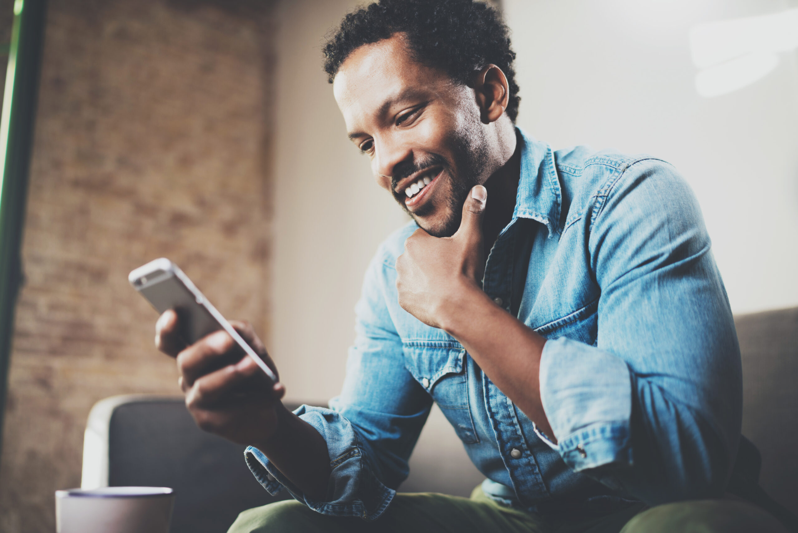 A man in a denim shirt sits on a couch, smiling and engaged with his smartphone, perhaps reading about employee retention strategies. With one hand on his chin and a coffee mug nearby, the blurred background suggests a cozy and thoughtful atmosphere.
