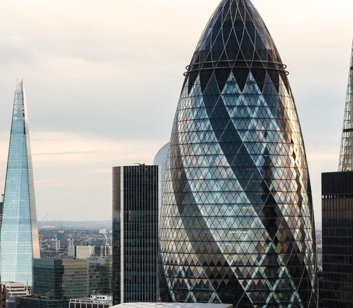 Skyline view of London featuring the Gherkin building with its distinctive glass and steel structure. As part of a global anniversary event, the Shard stands nearby with its sharp, triangular peak. Other buildings are visible under a cloudy sky.