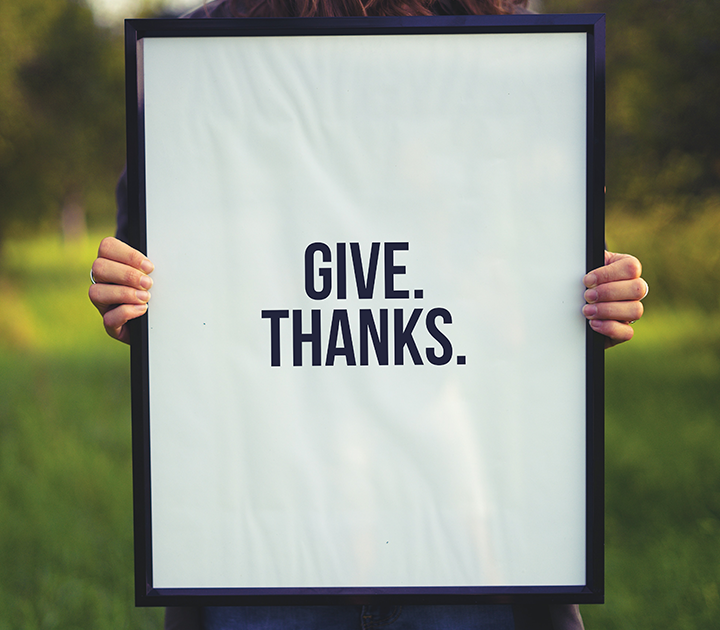 A person holds up a framed sign with the words GIVE. THANKS. in bold black letters, subtly reminding us of the importance of employee recognition. The background is a blurred green outdoor setting.