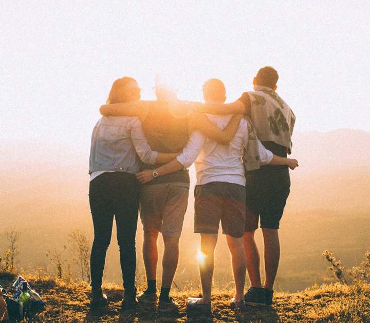 Four people stand together on a hilltop at sunset, arms around each other, looking at a scenic view. They are silhouetted by the sun, creating a warm, glowing atmosphere.