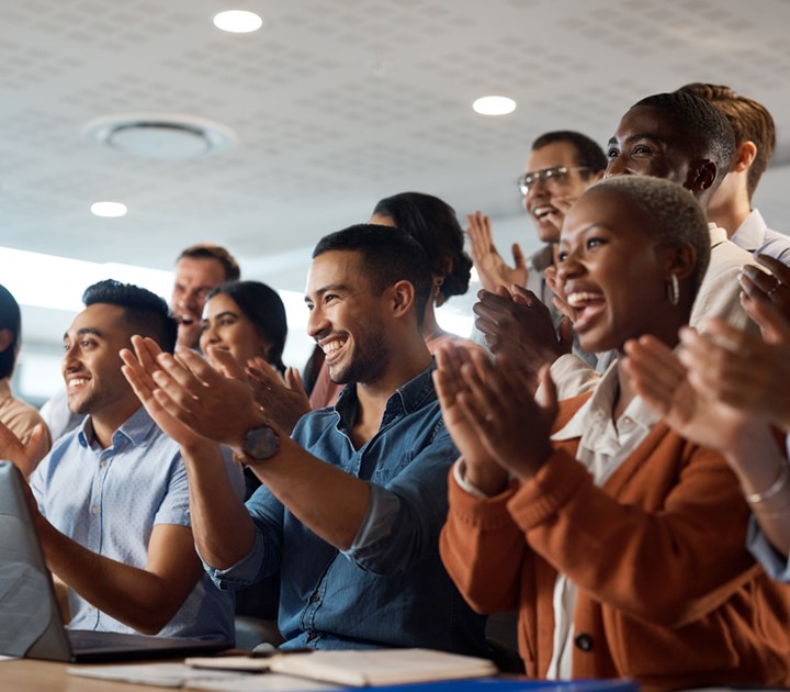 A diverse group of people in business attire are seated, facing forward, clapping and smiling in a show of peer recognition. The setting appears to be a lecture or conference room, with a ceiling and lights visible at the top.