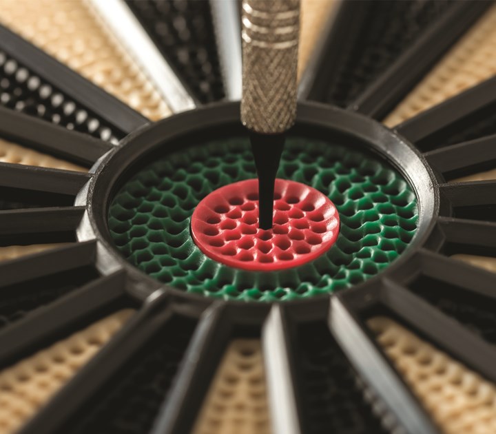 Extreme close-up of a dartboard with a dart hitting the red bullseye. The surrounding area is green with a textured pattern. The image captures the precision of the dart hitting the center.