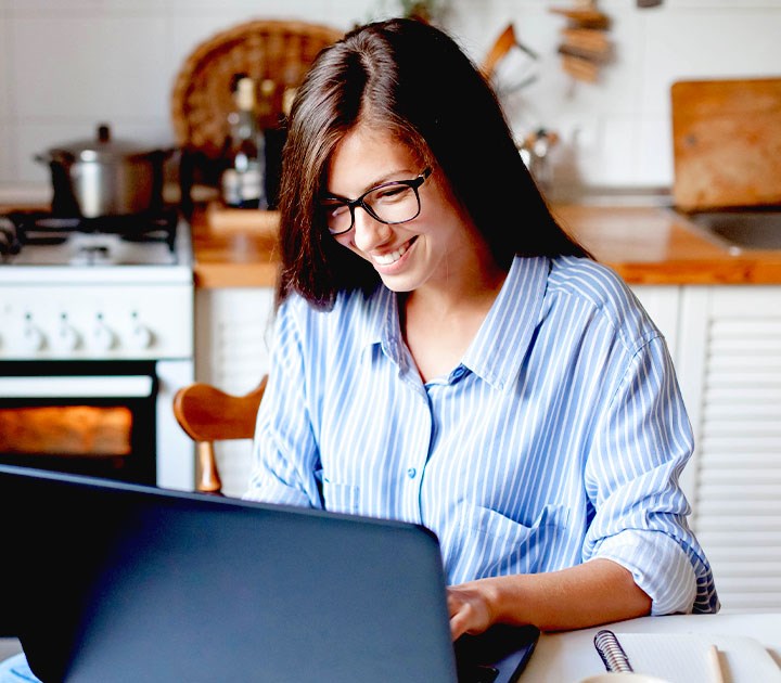 A person with long hair and glasses is smiling, driven by intrinsic motivation, as they work on a laptop in a cozy kitchen. They are wearing a striped shirt, and the background features a stove, wooden counter, and kitchen utensils.