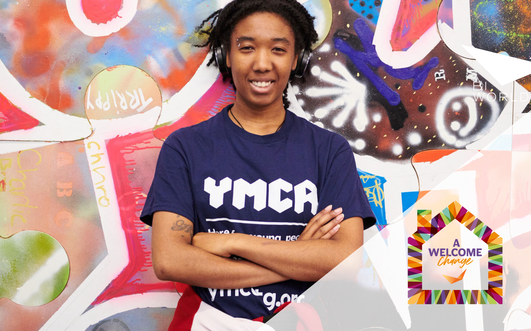 A person wearing a navy YMCA shirt stands with arms crossed, smiling in front of a colorful graffiti wall. In the corner, theres a graphic reading A Welcome Change in a house-shaped frame.