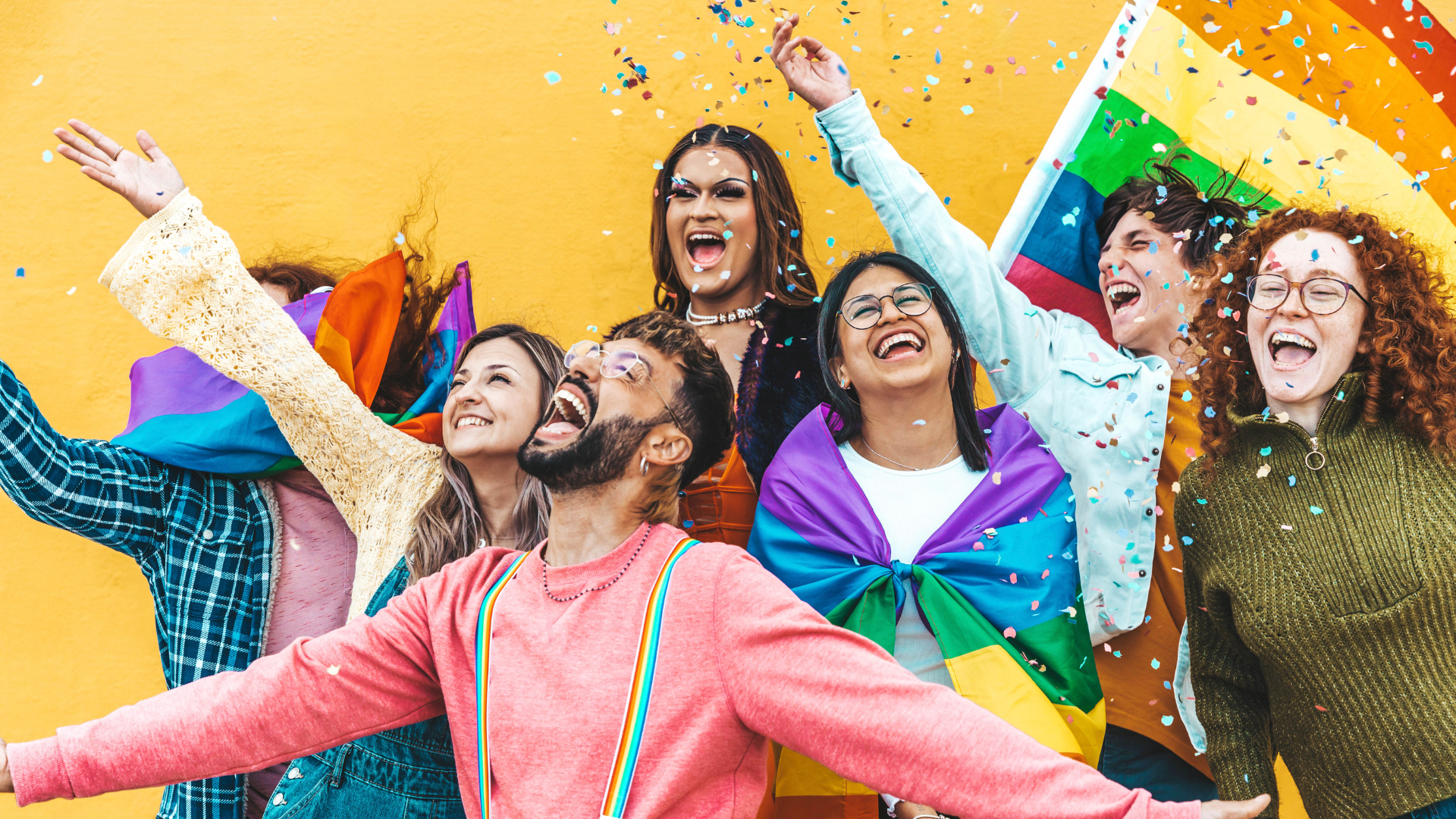 A diverse group of joyful people celebrate in front of a yellow wall, wearing rainbow flags, surrounded by confetti, and smiling, showing support for LGBTQ+ pride.