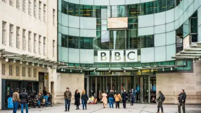 People stand and walk outside the glass entrance of the BBC Broadcasting House in London. The large BBC logo is prominently displayed above the entrance.