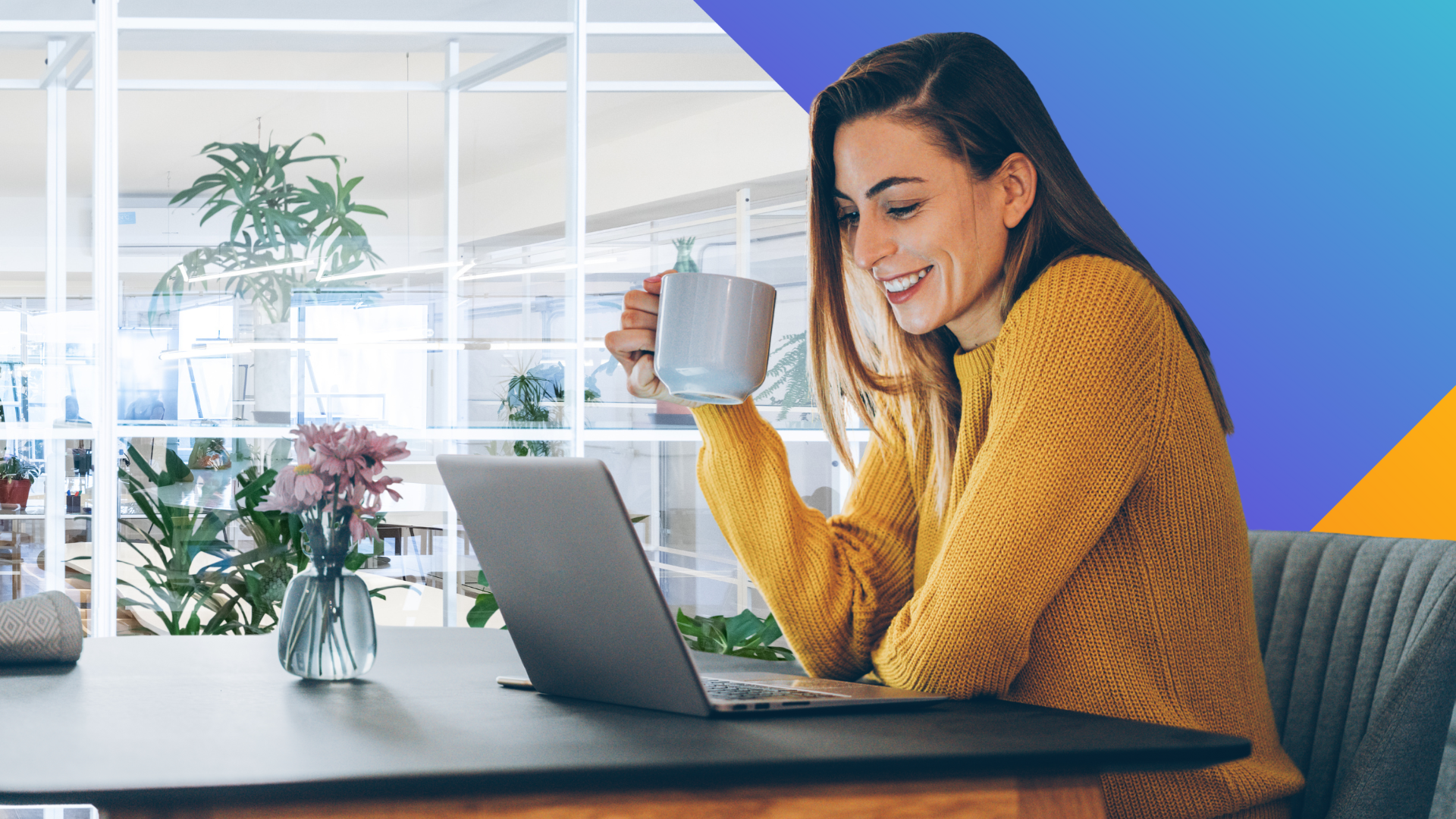 A woman in a yellow sweater sits at a table with a laptop, smiling and holding a mug. A vase with flowers is on the table, and the background shows a modern, bright office with plants and glass walls.