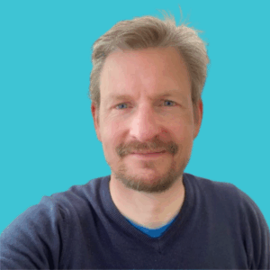 A man with light brown hair and a short beard smiles at the camera. He is wearing a dark blue sweater over a blue shirt, with a solid teal background behind him.