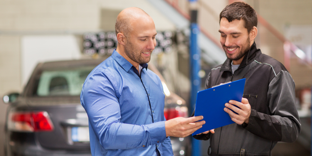 Two men smiling and discussing a document on a blue clipboard in a garage. One wears a blue shirt, and the other wears a black mechanics uniform. A car is in the background.