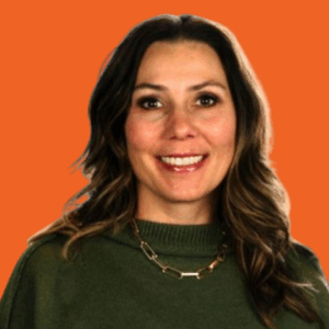 A woman with long brown hair, wearing a green sweater and a gold chain necklace, smiles in front of an orange background.