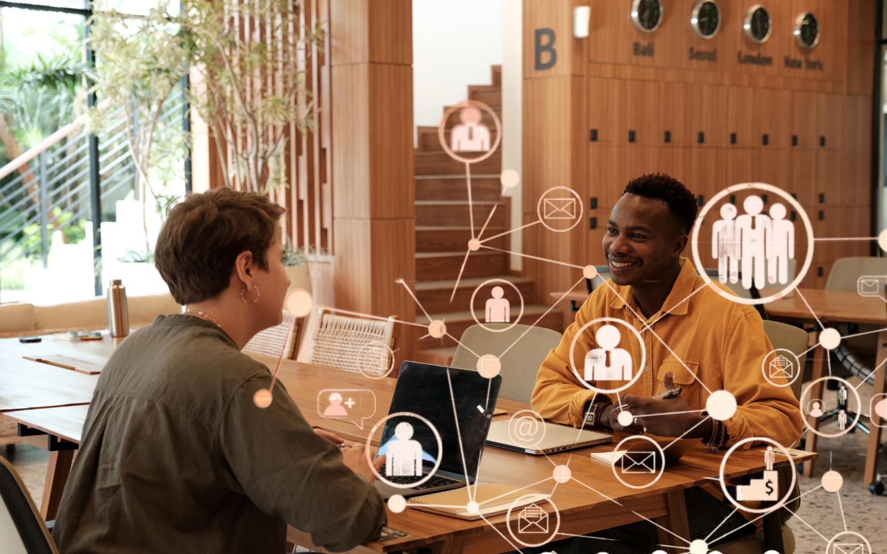 Two people sit across from each other at a table in a modern office, smiling and talking. Digital icons representing communication and networking are overlaid on the image.