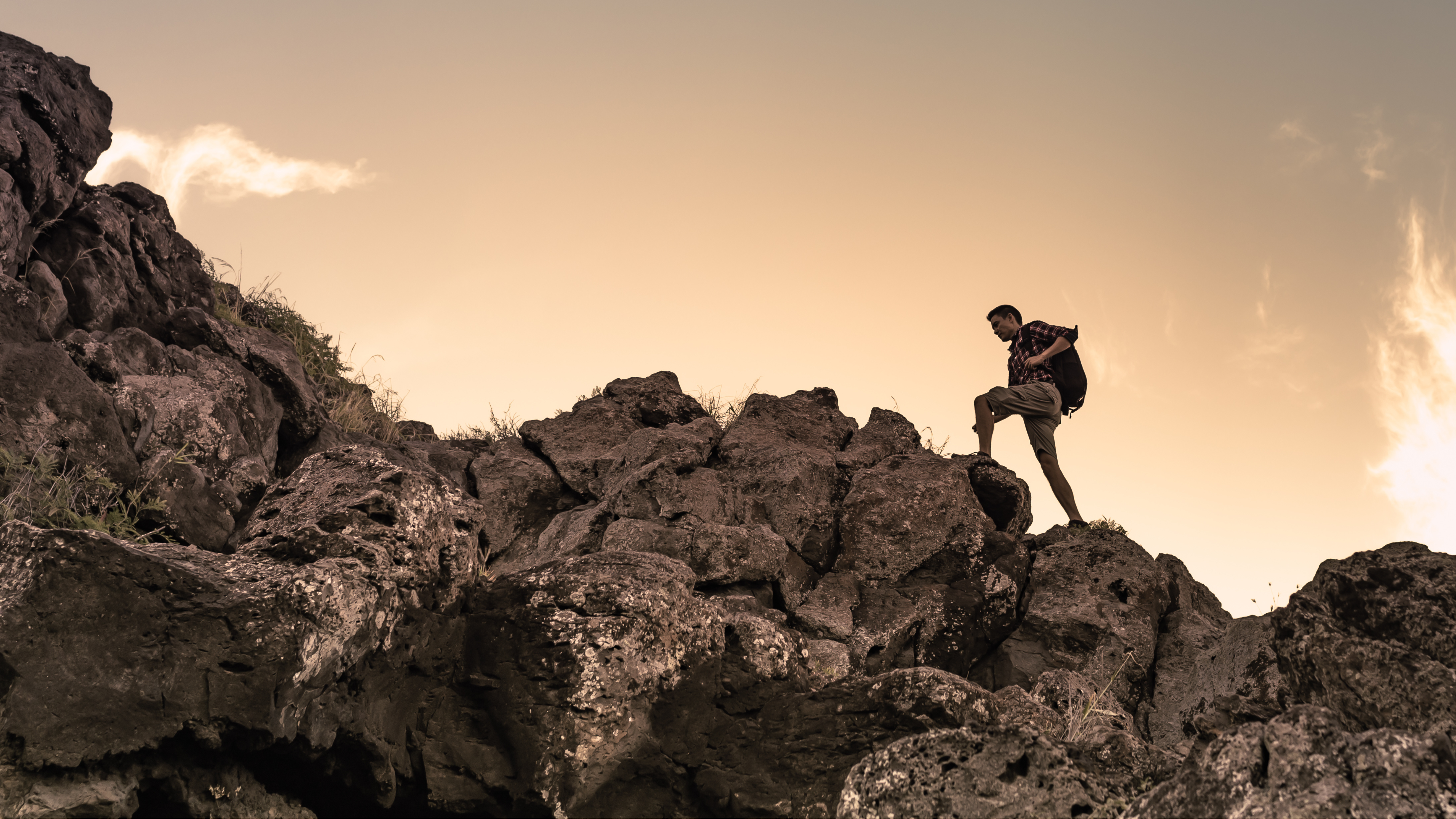 A person with a backpack climbs over large, rugged rocks at sunset, with a warm orange sky and wispy clouds in the background.