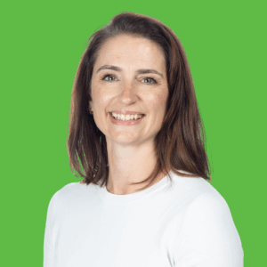 A woman with long brown hair wearing a white top smiles at the camera in front of a bright green background.