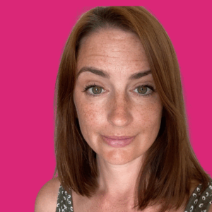A woman with straight, shoulder-length brown hair, light skin, and hazel eyes is smiling slightly. She is wearing a sleeveless top with a subtle pattern, and the background is a solid bright pink.