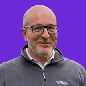 A smiling older man with glasses, short gray hair, and a beard, wearing a gray Regatta Great Outdoors jacket, stands in front of a solid purple background.