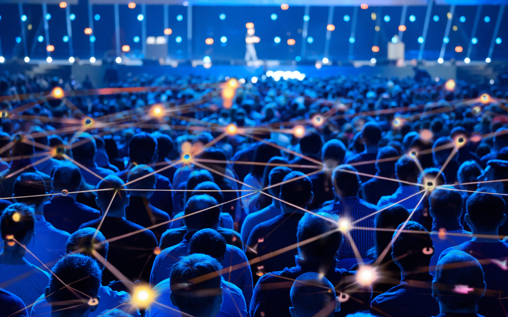 A large audience sits in a dimly lit auditorium facing a stage, where a speaker stands in front of a blue, illuminated backdrop. Abstract digital network lines and glowing nodes overlay the image, symbolizing connectivity.