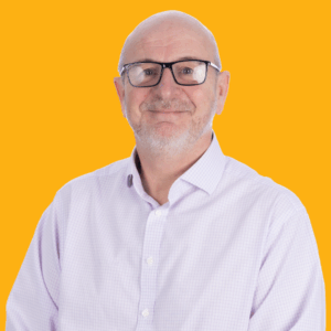 A smiling older man with glasses, a bald head, and a short white beard wearing a light purple shirt, standing in front of a solid bright orange background.