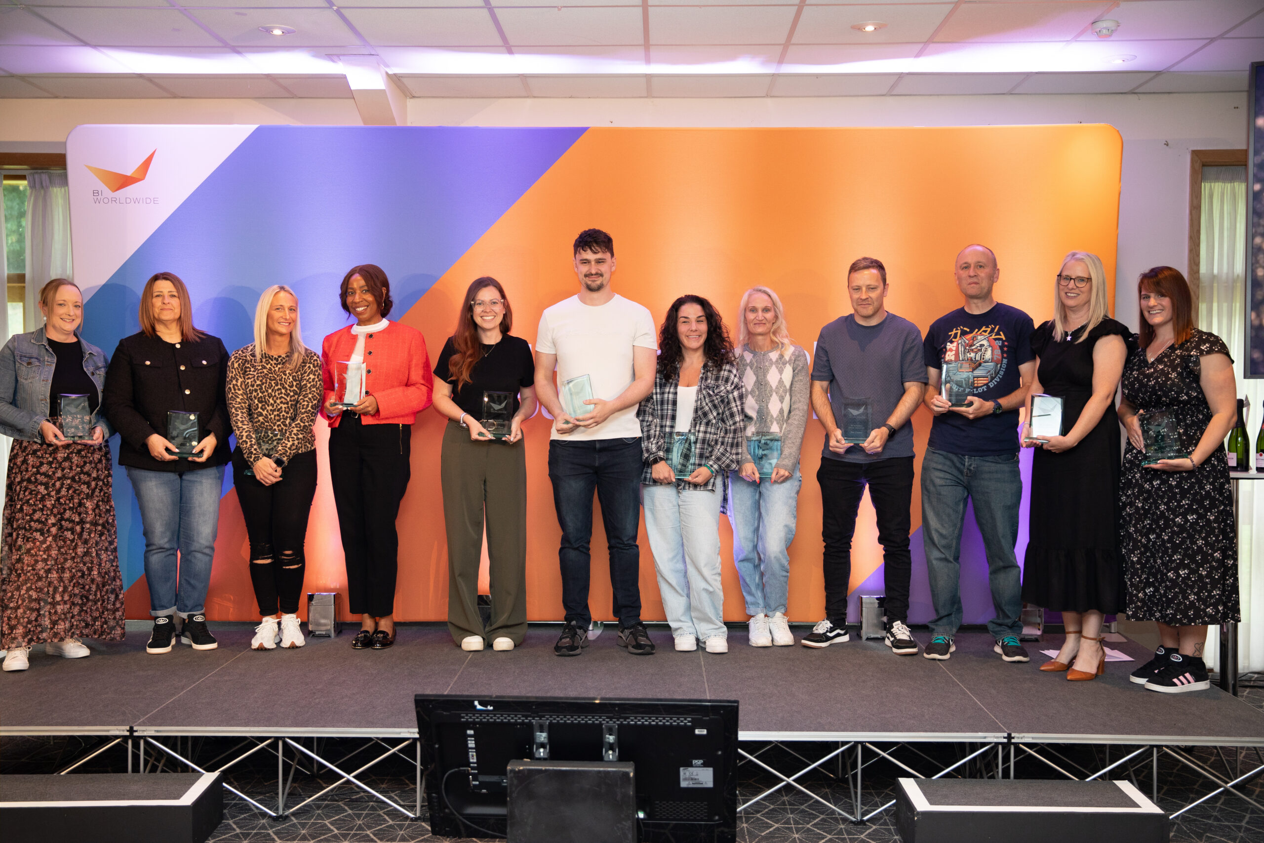 Twelve people stand side by side on a stage, holding glass awards and smiling, in front of an orange and blue backdrop. The group consists of men and women dressed in casual to semi-formal attire.