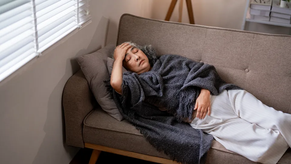 An older woman with gray hair lies on a couch, covered in a dark blanket, resting her head on a pillow and holding her forehead, appearing tired or unwell. Light streams in through nearby window blinds.