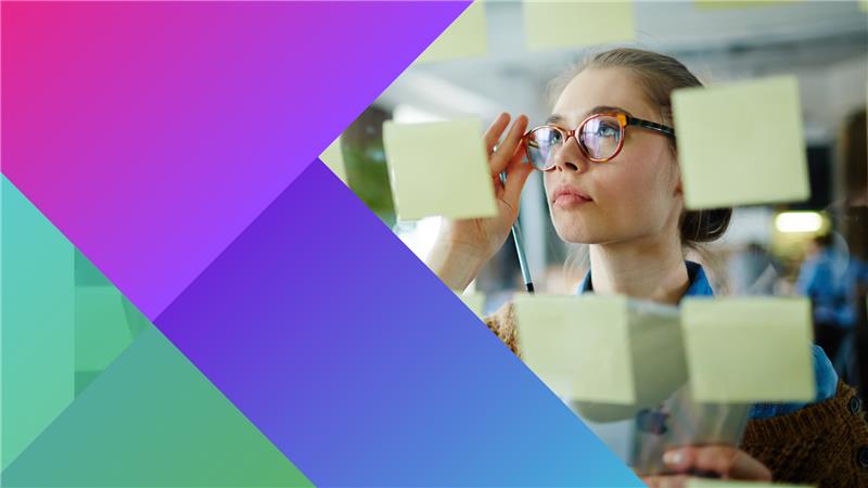 A woman wearing glasses looks thoughtfully at sticky notes on a glass wall, holding a pen. Colourful geometric shapes overlay the left side of the image.