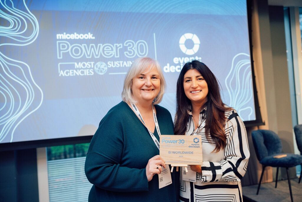 Two women smiling and holding a Power 30 Most Sustainable Agencies award plaque. A presentation screen behind them displays “micebook Power 30 Most Sustainable Agencies” and “event decision.”.