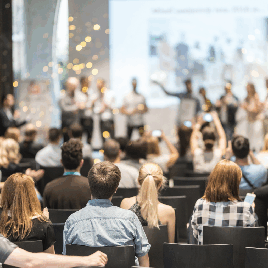 Audience sitting in rows of chairs facing a stage where people are standing, some holding objects. Several attendees are taking photos or videos with their phones. The atmosphere appears lively, with blurred lights in the background.