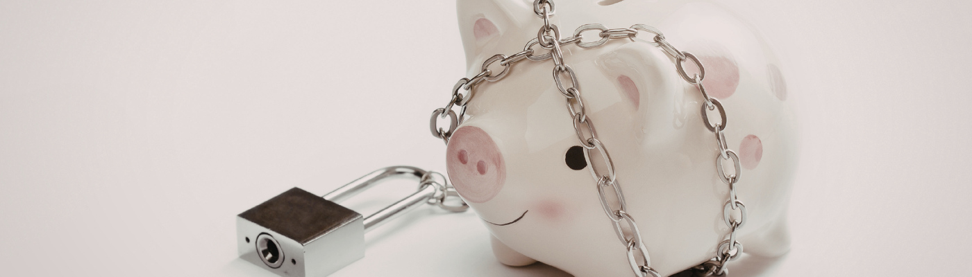 A white ceramic piggy bank with pink cheecks and polka dots is wrapped in a metal chain secured with a padlock, symbolizing financial security or restricted access to savings.