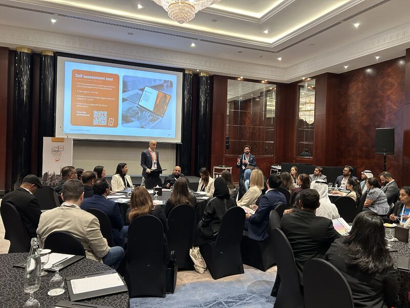 A group of people in business attire sit at round tables in a conference room, listening to a speaker who stands near a screen displaying a Self assessment tool slide. The setting is modern and formal.