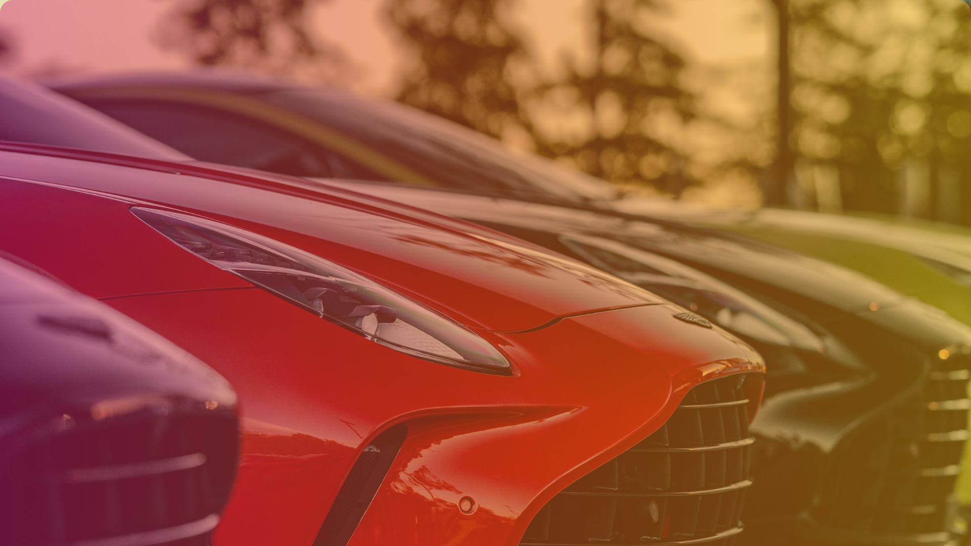 Close-up view of the front ends of several luxury sports cars parked in a row, with a prominent bright red car in the foreground and a warm, gradient filter overlaying the image.