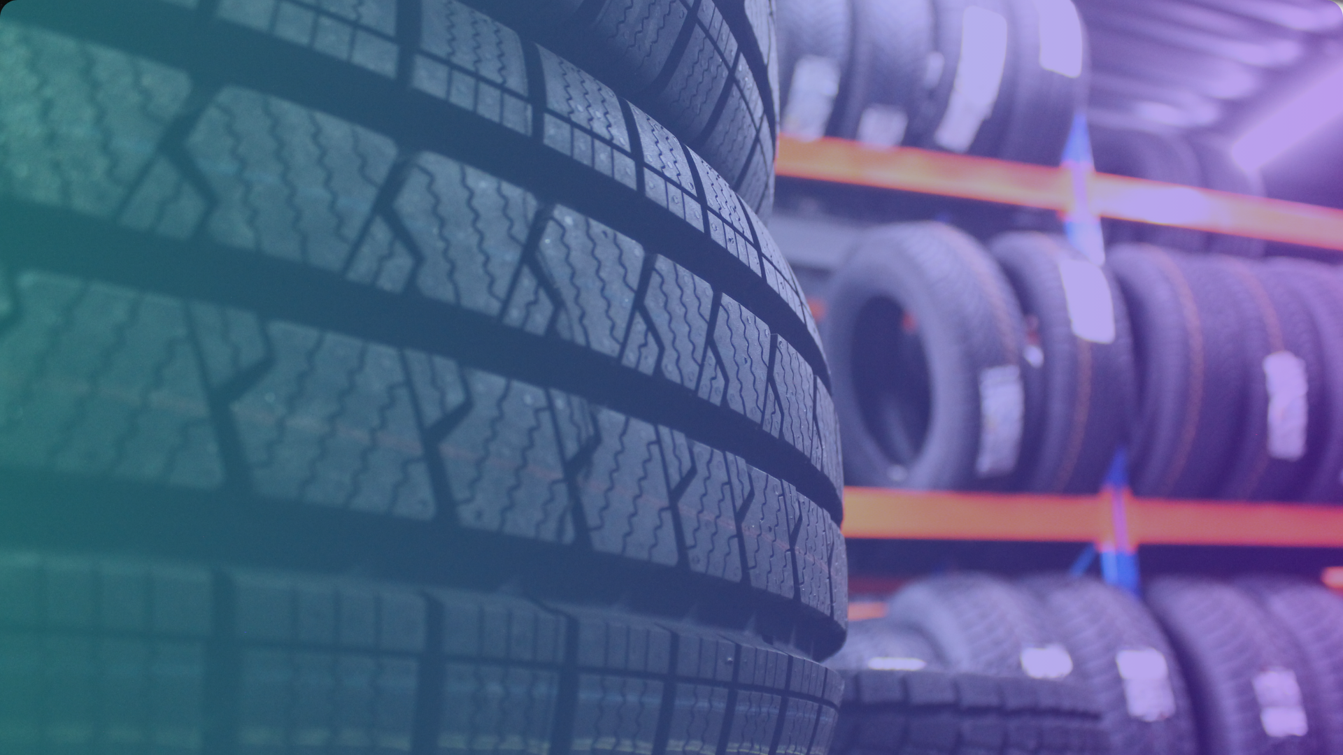Close-up of stacked car tires with visible tread patterns in the foreground, and more tires arranged on orange shelves in the background, inside a tire shop or storage area.