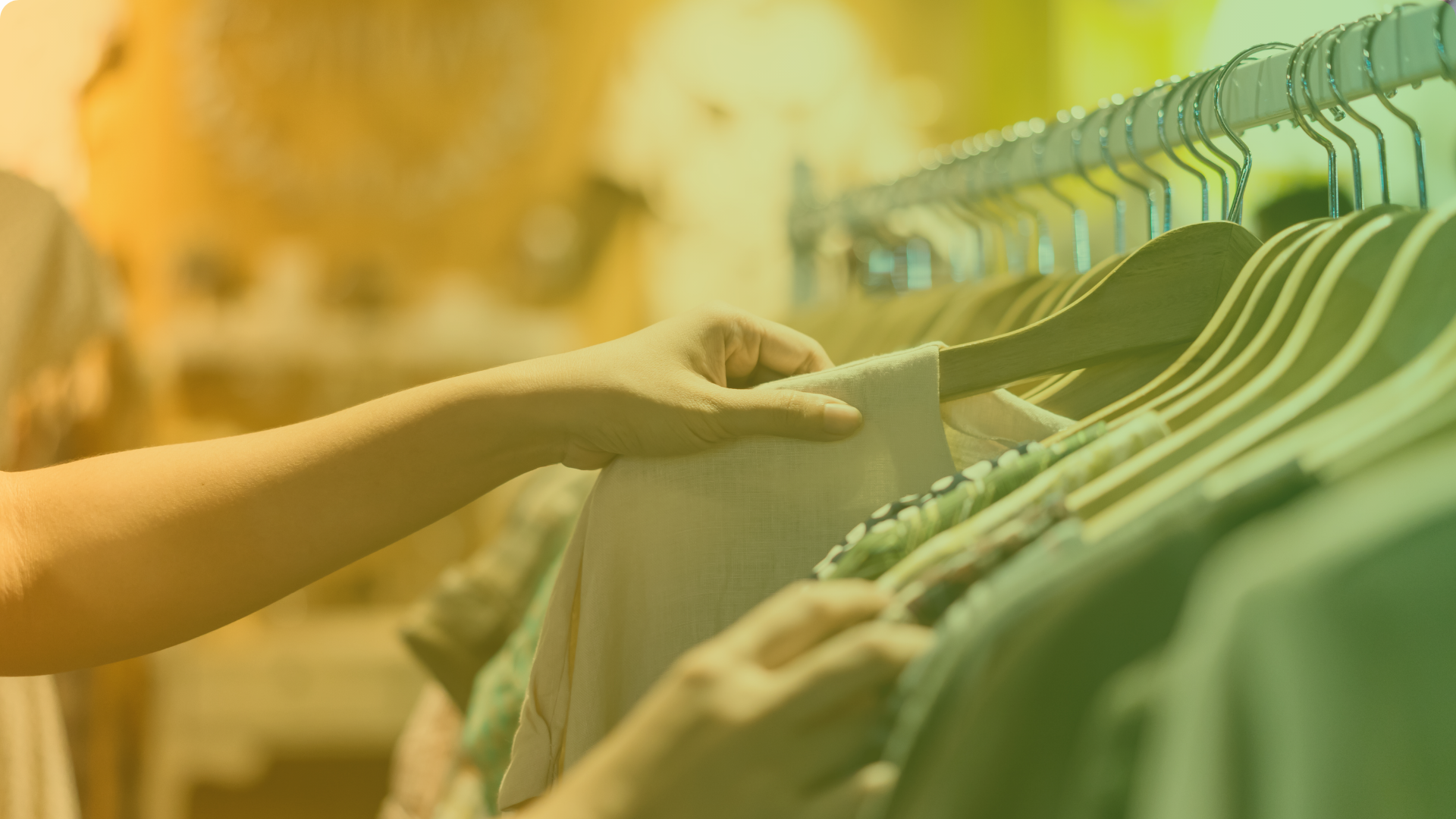 A person’s hand browses through clothes on hangers in a store, selecting a light-colored shirt from a rack of assorted garments.