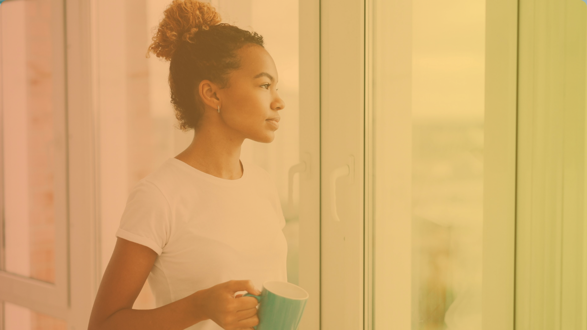 A woman with curly hair tied up, wearing a white t-shirt, holds a blue mug and gazes thoughtfully out of a large window. The image has a warm, yellowish tint.