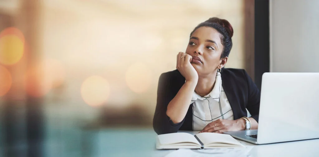 A woman sits at a desk with an open notebook and a laptop, resting her chin on her hand and looking thoughtfully out of frame. She appears to be deep in thought or daydreaming.