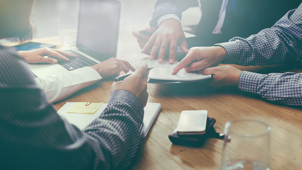 Four people sit around a wooden table working together. They have a laptop, notepads, and documents. One person points at a piece of paper while others take notes. Sunlight streams onto the table.