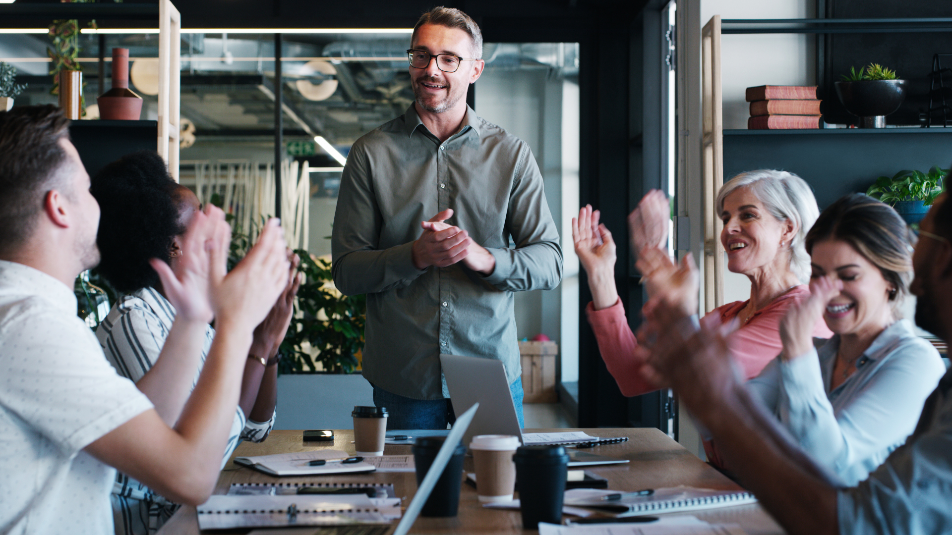 A man stands and smiles while colleagues seated around a conference table applaud him. The group appears happy and engaged in a modern, well-lit office setting with laptops and notebooks on the table.