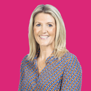 A smiling woman with straight blonde hair, wearing a patterned blue and orange blouse, poses in front of a bright pink background.