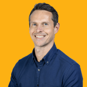 A smiling man with short brown hair wearing a dark blue button-up shirt stands in front of a solid yellow background.