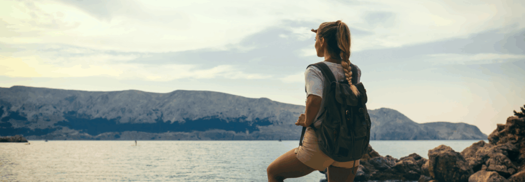 A woman with a backpack stands on rocky terrain by the water, looking at distant mountains under a cloudy sky, enjoying a scenic outdoor view.