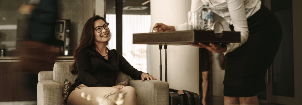 A woman sitting on a chair in an airport lounge smiles as a staff member serves her a drink on a tray. There is luggage beside her and people walking in the background.