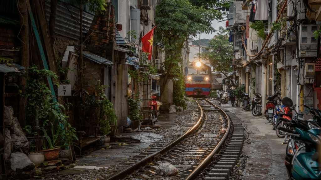 A train approaches on narrow tracks running through a residential alley lined with plants, motorbikes, and closely packed buildings with balconies and laundry hanging outside.