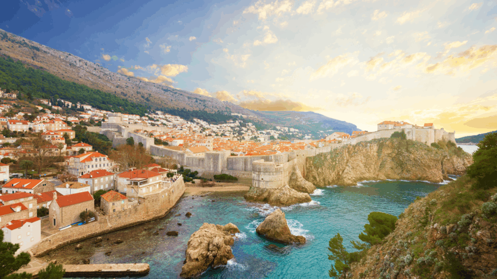 A scenic view of Dubrovnik, Croatia, showing the historic city walls, red-roofed buildings, and rocky coastline at sunset, with the hills rising in the background.