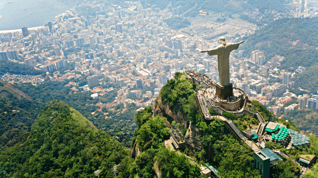 Aerial view of the Christ the Redeemer statue atop a green mountain with the city of Rio de Janeiro, Brazil, spread out below.