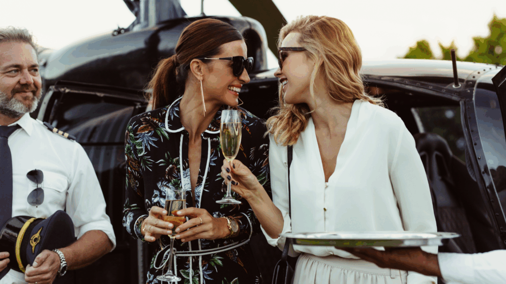 Two women in stylish outfits laugh and toast with champagne beside a helicopter, while a pilot in uniform stands nearby, holding his hat and smiling.