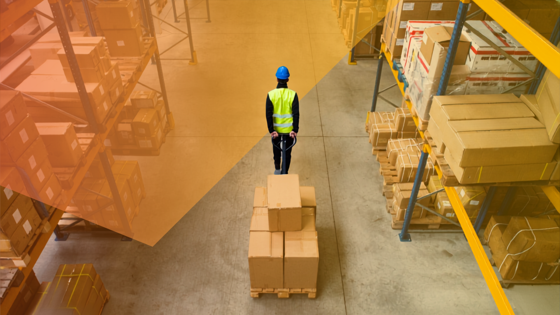 A worker in a yellow vest and blue helmet moves a pallet of cardboard boxes through a large warehouse with metal shelves stocked with more boxes.