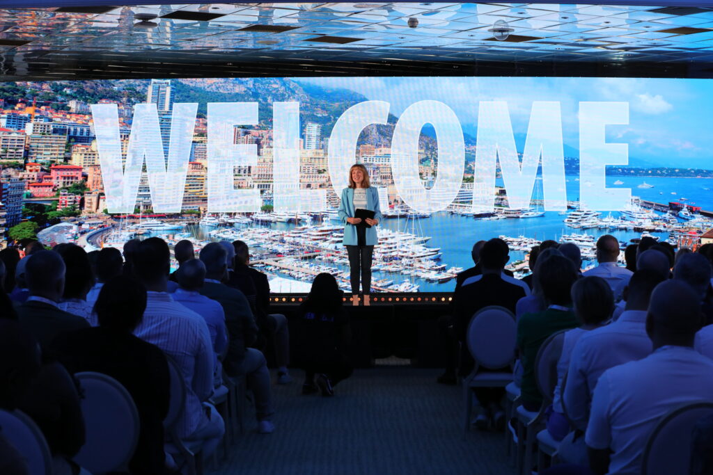 A woman stands on stage addressing an audience, with a large screen behind her displaying WELCOME over an image of a coastal city and marina.
