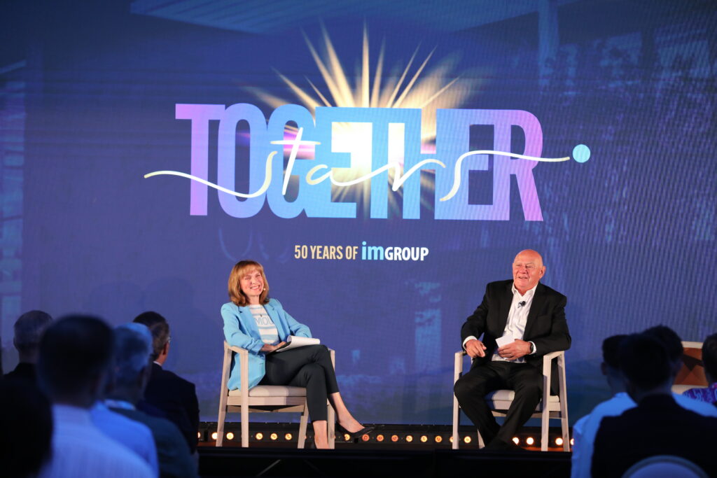 Two people sit on stage, smiling, with a colourful TOGETHER and 50 YEARS OF imGROUP sign behind them. Audience members are visible in the foreground, watching the event.