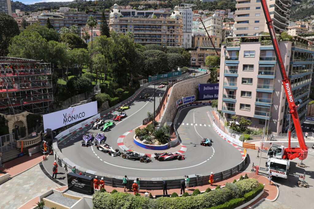 Formula E racing cars navigate a tight corner on a city street circuit in Monaco, with grandstands, buildings, and spectators visible in the background. Race marshals and a crane are also present near the track.