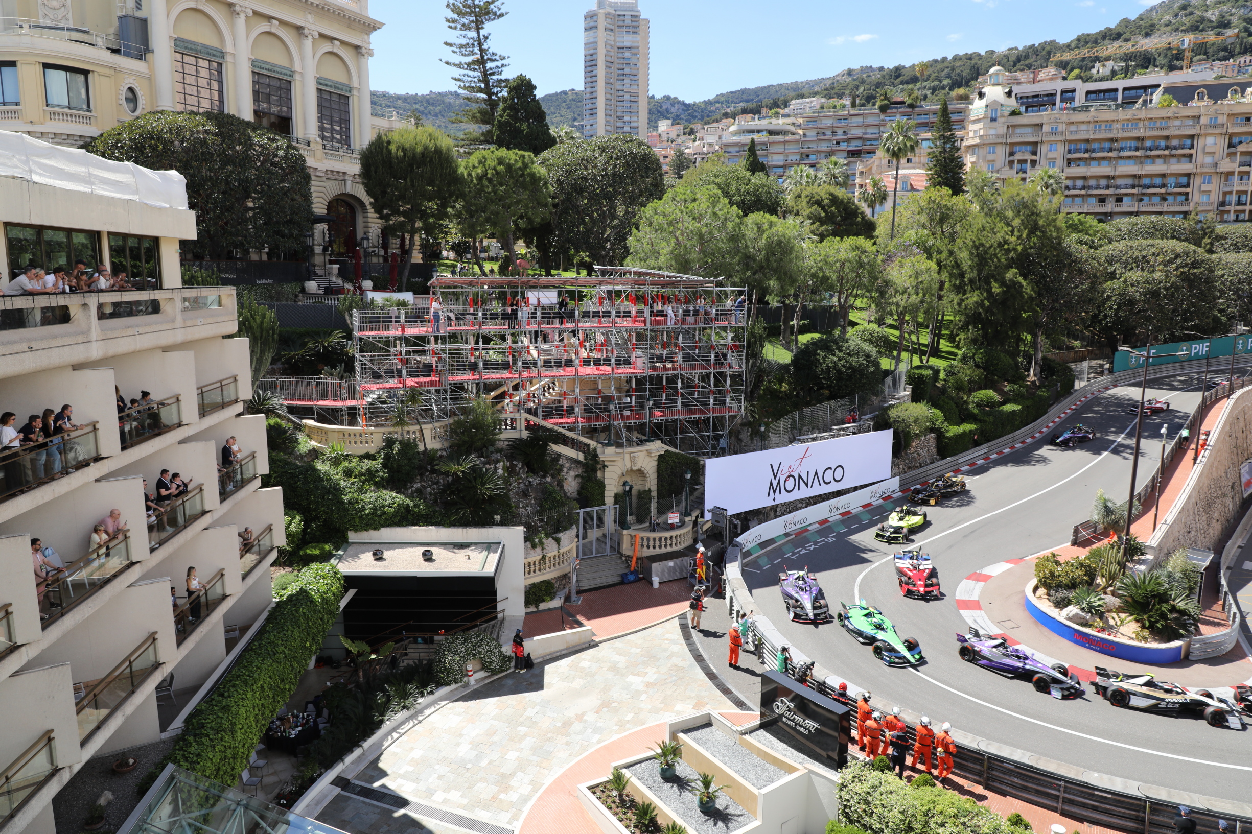 Formula 1 cars race round a tight corner in Monaco, with spectators watching from balconies and grandstands surrounded by lush greenery and tall buildings in the background.