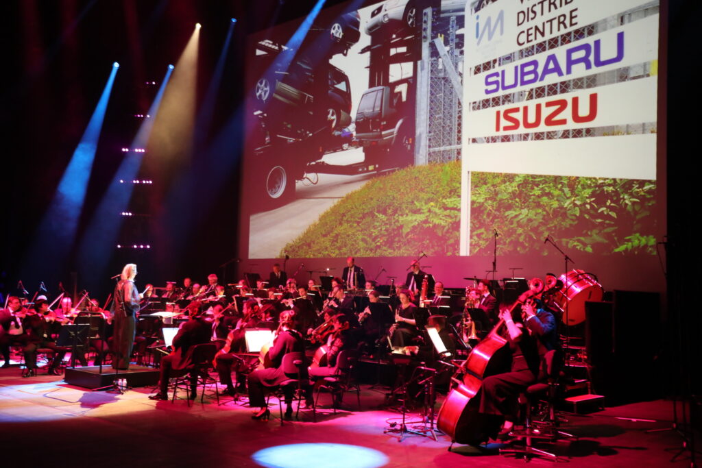 An orchestra performs onstage with a conductor, with a large projection behind them showing cars and logos for Subaru and Isuzu. The stage is illuminated with dramatic lighting.